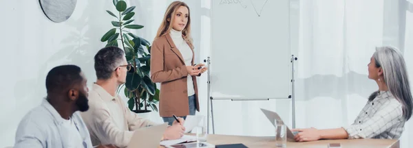 Plan panoramique d'une femme d'affaires debout près d'un tableau à feuilles mobiles et parlant avec des collègues multiculturels — Photo de stock
