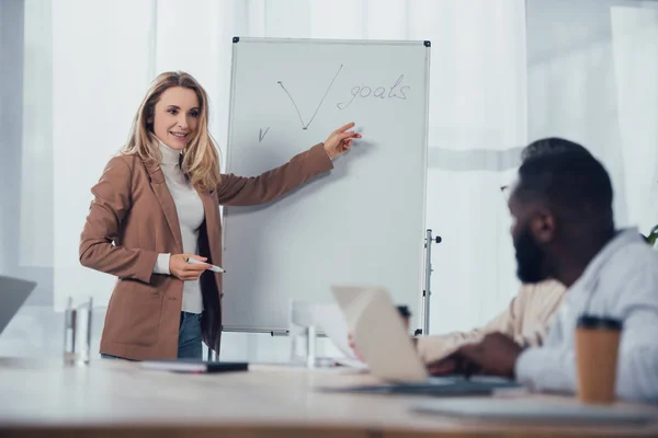 Selective focus of smiling businesswoman pointing with finger at goals lettering and talking with african american colleague — Stock Photo