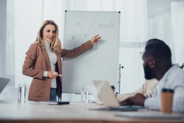 Focalizzazione selettiva di una donna d'affari sorridente che punta il dito sugli obiettivi lettering e parla con un collega afroamericano — Foto stock