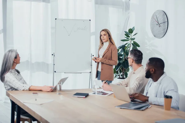 Geschäftsfrau steht neben Flipchart mit Zielbuchstaben und spricht mit multikulturellen Kollegen — Stockfoto