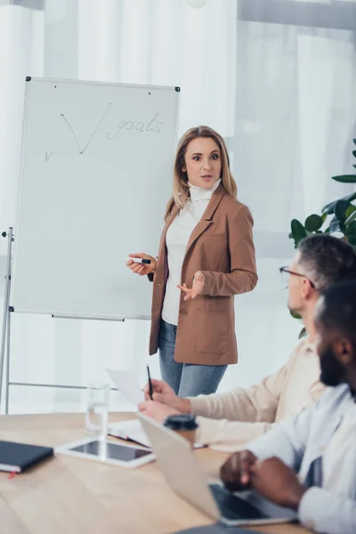 Enfoque selectivo de la mujer de negocios de pie cerca del rotafolio con letras de objetivos y hablar con colegas multiculturales - foto de stock