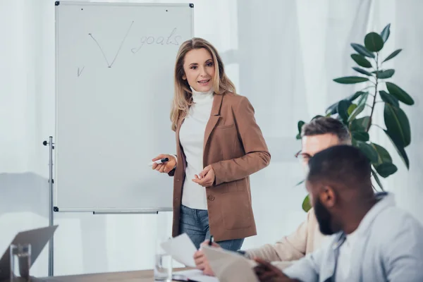 Enfoque selectivo de la mujer de negocios conmocionada de pie cerca del rotafolio con letras de objetivos y hablar con colegas multiculturales - foto de stock