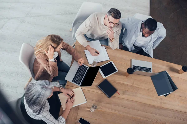 Visión general de colegas multiculturales conversando y sentándose a la mesa durante la reunión - foto de stock