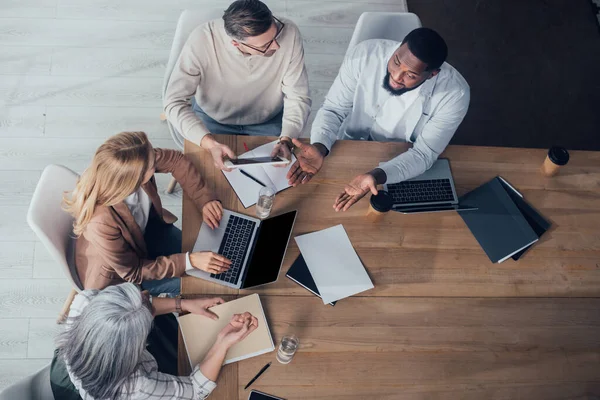 Visión general de colegas multiculturales conversando y sentándose a la mesa durante la reunión - foto de stock