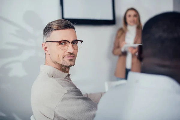 Enfoque selectivo de sonriente hombre de negocios mirando a la cámara y sentado cerca de un colega afroamericano - foto de stock