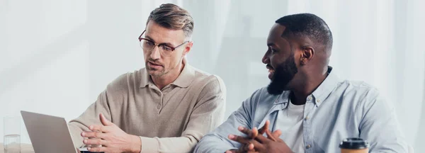 Panoramic shot of businessman pointing with hand at laptop and talking with smiling african american colleague — Stock Photo