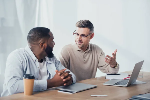 Smiling multicultural colleagues sitting at table and talking in creative agency — Stock Photo