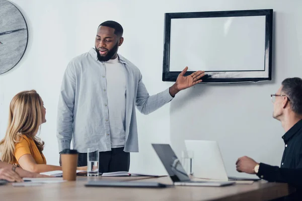 Sorridente empresário afro-americano apontando com a mão na tv e conversando com colegas — Fotografia de Stock