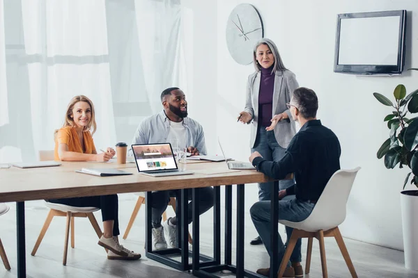 KYIV, UCRANIA - 6 DE DICIEMBRE DE 2019: sonrientes colegas multiculturales hablando con una mujer de negocios asiática cerca de la computadora portátil con el sitio web shutterstock - foto de stock