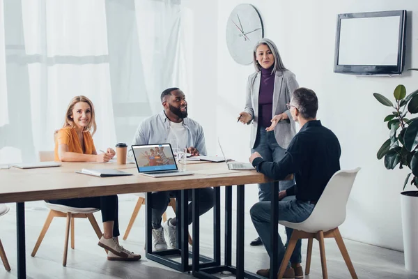 KYIV, UCRANIA - 6 DE DICIEMBRE DE 2019: sonrientes colegas multiculturales hablando con la mujer de negocios asiática cerca de la computadora portátil con el sitio web couchsurfing - foto de stock