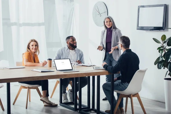 KYIV, UCRANIA - 6 DE DICIEMBRE DE 2019: sonrientes colegas multiculturales hablando con la mujer de negocios asiática cerca de la computadora portátil con google en la pantalla - foto de stock