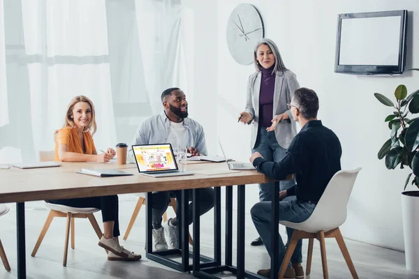 KYIV, UCRANIA - 6 DE DICIEMBRE DE 2019: sonrientes colegas multiculturales hablando con la mujer de negocios asiática cerca de la computadora portátil con el sitio web aliexpress - foto de stock