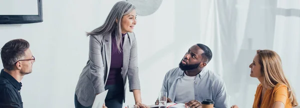 Plano panorámico de sonriente mujer de negocios asiática hablando con colegas multiculturales en la oficina - foto de stock