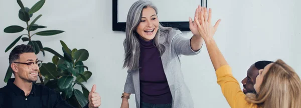 Panoramic shot of smiling multicultural colleagues giving high five and showing like — Stock Photo