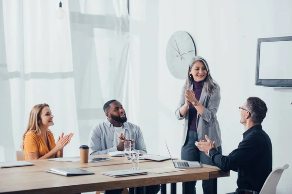 Sonriente multicultural colegas aplaudiendo y mirando asiático mujer de negocios - foto de stock