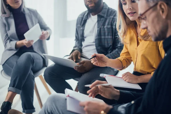 Vista recortada de colegas multiculturales con gadgets sentados y hablando en la oficina - foto de stock