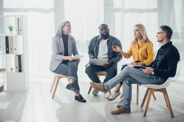 Colegas multiculturales con aparatos sentados y hablando en la oficina - foto de stock