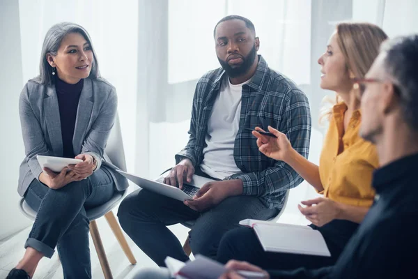 Focus selettivo di colleghi multiculturali con gadget seduti e che parlano in ufficio — Foto stock