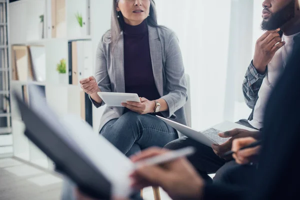 Cropped view of multicultural colleagues with gadgets sitting and talking in office — Stock Photo