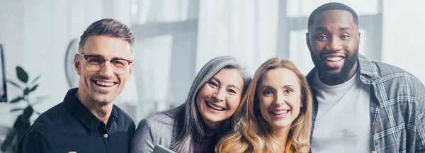 Panoramic shot of smiling multicultural colleagues looking at camera — Stock Photo