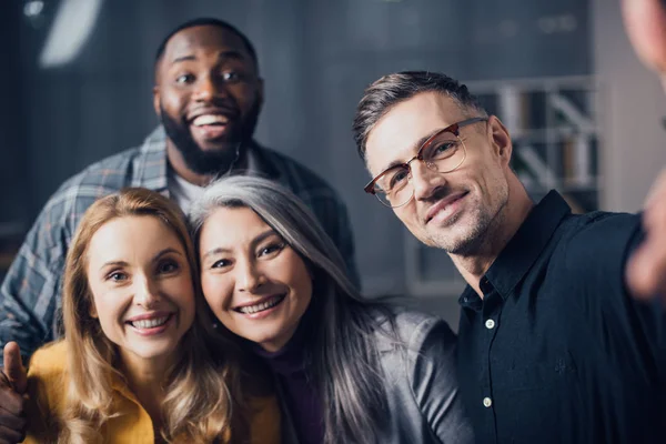 Selektiver Fokus lächelnder multikultureller Kollegen, die in die Kamera schauen und Selfies machen — Stockfoto