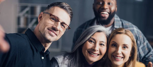 Plano panorámico de sonrientes colegas multiculturales mirando a la cámara y tomando selfie - foto de stock