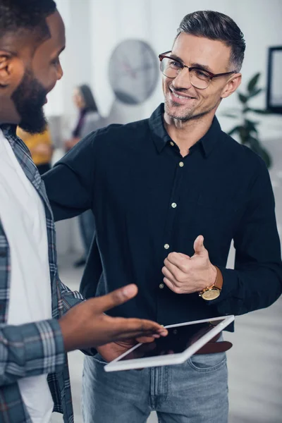 Cropped view of smiling african american businessman pointing with hand at digital tablet and his colleagues showing like — Stock Photo