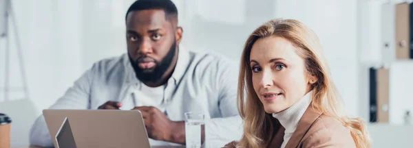 Panoramic shot of multicultural colleagues sitting at table and looking at camera — Stock Photo