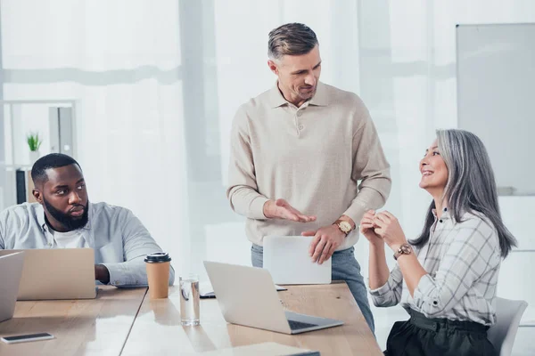 Colegas multiculturales sonriendo y hablando en agencia creativa - foto de stock