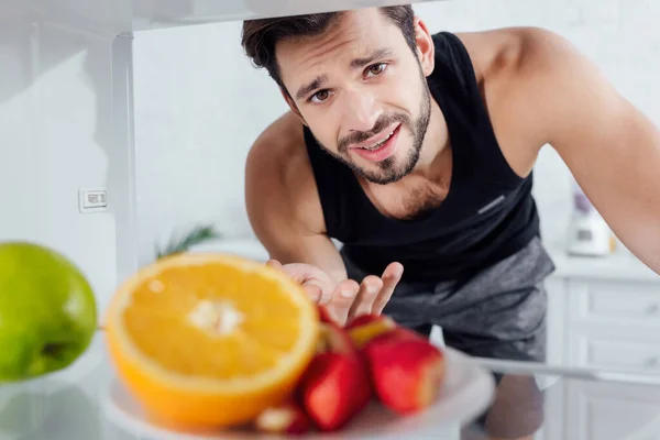 Selective focus of confused man looking at camera and gesturing near fruits in fridge — Stock Photo