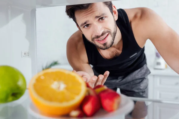 Enfoque selectivo del hombre confundido mirando a la cámara y haciendo gestos cerca de las frutas en la nevera - foto de stock