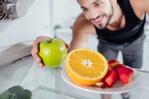 Selektiver Fokus des glücklichen Mannes, der Apfel aus dem Kühlschrank nimmt — Stockfoto