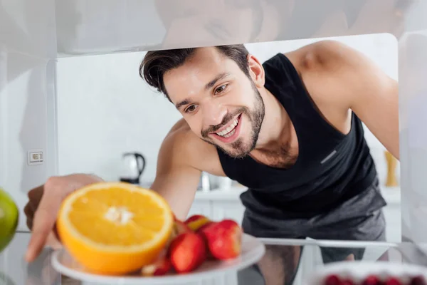 Messa a fuoco selettiva dell'uomo allegro che prende il piatto con frutta dal frigorifero — Foto stock