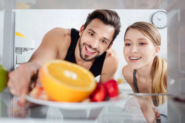 Selective focus of happy man and woman looking at tasty fruits in fridge — Stock Photo