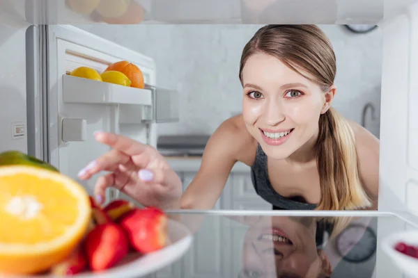 Focalizzazione selettiva di donna allegra che raggiunge frutta in frigorifero — Foto stock
