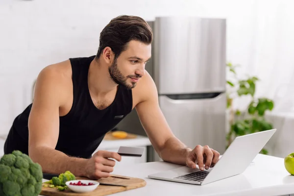 Homem bonito segurando cartão de crédito ao usar laptop na cozinha — Fotografia de Stock