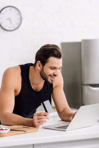Hombre feliz celebración de la tarjeta de crédito mientras se utiliza el ordenador portátil en la cocina - foto de stock