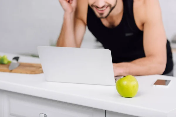 Vista recortada de hombre feliz utilizando el ordenador portátil cerca de manzana en la cocina - foto de stock