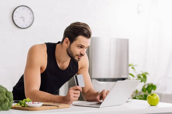Schöner Mann mit Kreditkarte, während er Laptop in der Nähe von Apfel und Brokkoli benutzt — Stockfoto