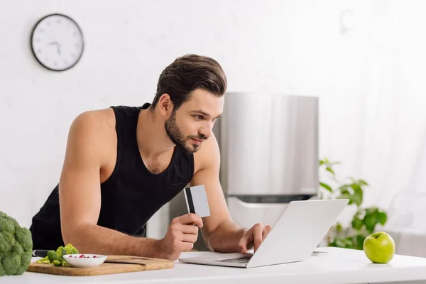 Hombre guapo celebración de la tarjeta de crédito mientras se utiliza el ordenador portátil cerca de manzana y brócoli — Stock Photo