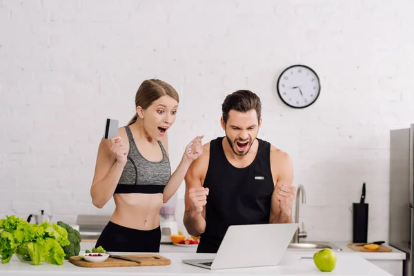 Hombre y mujer emocionados con tarjeta de crédito mirando portátil en la cocina - foto de stock