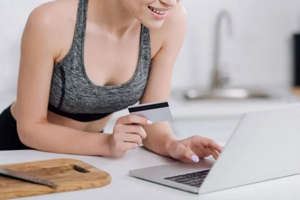 Vista cortada de menina alegre segurando cartão de crédito perto de laptop na cozinha — Fotografia de Stock