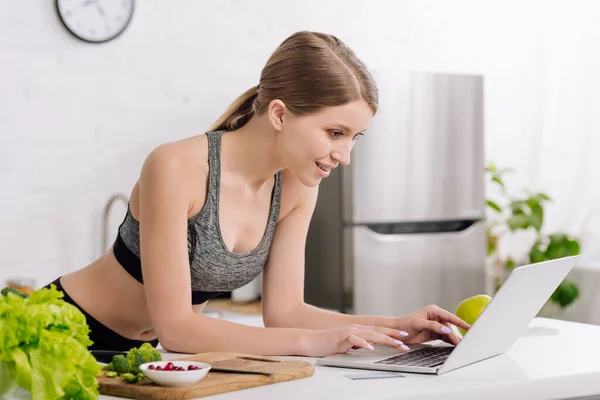 Cheerful girl typing on laptop keyboard near credit card and food in kitchen — Stock Photo