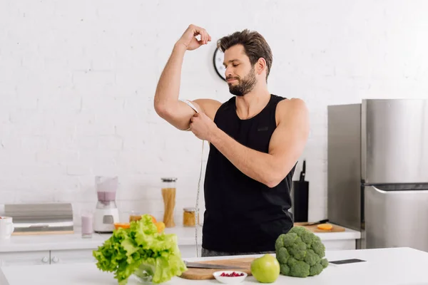 Homem bonito e esportivo que mede o músculo na mão perto de alimentos frescos — Fotografia de Stock