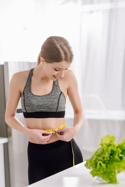 Fröhliches Mädchen misst Taille in der Nähe von frischem Salat in Küche — Stockfoto