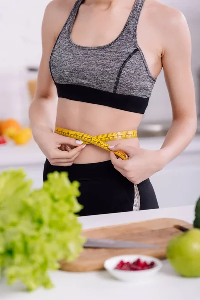 Vista recortada de niña deportiva que mide la cintura cerca de alimentos frescos en la cocina - foto de stock