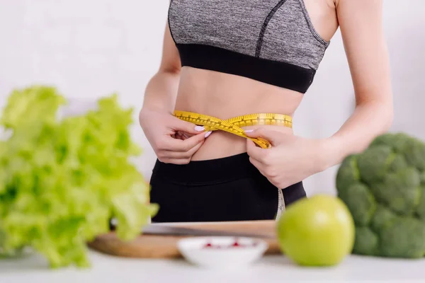 Vista cortada de menina esportiva medindo cintura perto de alimentos frescos — Fotografia de Stock