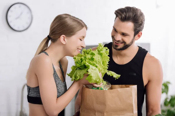 Веселая девушка держит салат возле бумажного пакета и счастливый бородатый мужчина на кухне — стоковое фото