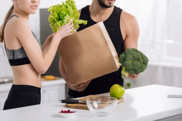 Vista recortada de la mujer tomando lechuga de la bolsa de papel cerca del hombre barbudo en la cocina - foto de stock