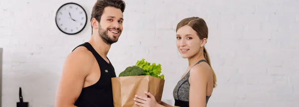 Panoramic shot of cheerful girl and happy man near paper bag with groceries — Stock Photo